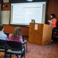 Image of a student in front of a podium presenting in front of an audience with a screen about computational linguistics in the background.