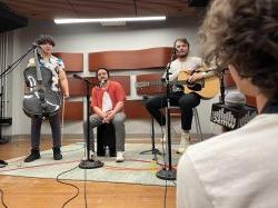 A person in the foreground is interviewing a three-piece band, two of which are holding intstruments.